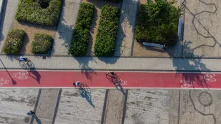 Rowerzyści na czerwonym pasie drogi rowerowej w otoczeniu zieleni.
