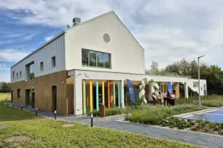 Nowoczesny budynek z dużymi oknami, otoczony zielenią i kolorowymi detalami.