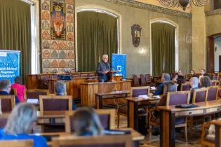 Konferencja w sali obrad z prelegentem i uczestnikami siedzącymi w rzędach.