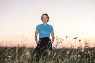 Kobieta w niebieskiej bluzce stoi w polu z wysoką trawą, w tle zachód słońca.