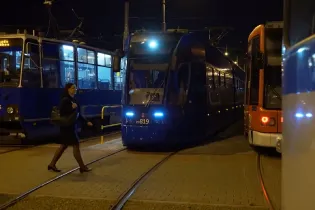 Tramwaj w nocy, kobieta przechodzi obok, inne pojazdy szynowe w tle.