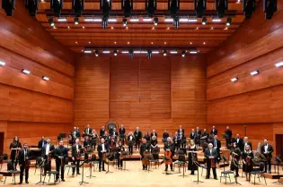 Orkiestra w sali koncertowej, muzycy w strojach formalnych, instrumenty gotowe do występu.