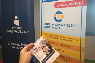 Zdjęcie przedstawia materiały promocyjne Centrum Obywatelskiego Centrum C 10 i Instytutu Polityk Publicznych. Na pierwszym planie dłoń trzyma ulotkę targów pracy, a w tle stoją banery z logo i ofertą 