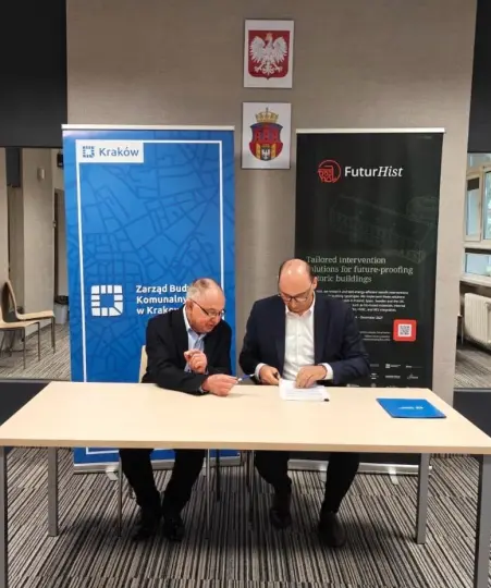 Dwaj mężczyźni podpisują dokumenty przy stole, w tle flagi i logotypy instytucji.