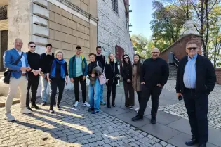 Grupa ludzi na tle historycznego budynku i drzew w słoneczny dzień.