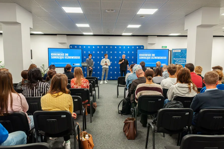 zdjęcia z rozpoczęcia KDSU 2026, 3 osoby przemawiają na tle ekranów i ścianki z logotypami miasta Krakowa, na pierwszym planie rzędy uczniów i nauczycieli