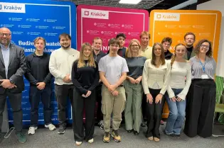 Na tle kolorowych plansz pozuje do wspólnej fotografii grupa uśmiechniętych osób. Zdecydowana większość z nich to młodzież.   