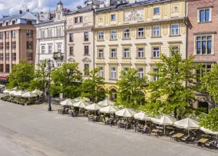 Kawiarnie z parasolami na placu, otoczone kamienicami i zielonymi drzewami.