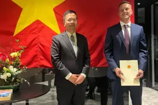 W nowoczesnym pomieszczeniu biurowym na tle flagi Wietnamu stoi dwóch mężczyzn niższy z nich po lewej Ambasador Wietnamu w Polsce a po prawej nowo mianowany konsul tego kraju trzymający teczkę