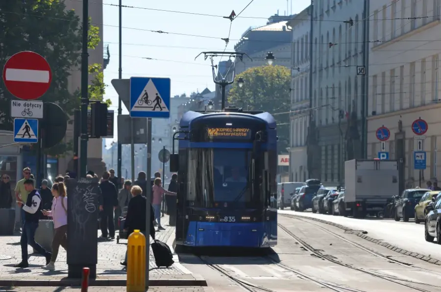 Niebieski tramwaj na ulicy, przechodnie i znaki drogowe w tle.