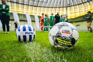 Dwa piłki nożne na murawie stadionu, w tle zawodnicy w strojach sportowych.