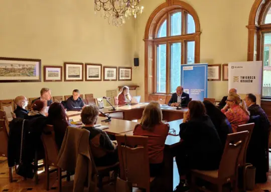 Spotkanie grupy ludzi w sali konferencyjnej, przy stole, z dokumentami i prezentacjami.