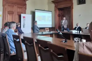 Prezentacja w sali konferencyjnej z uczestnikami siedzącymi przy stole.