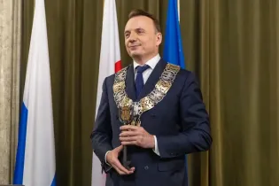 Mężczyzna w garniturze z łańcuchem prezydenckim, trzymający berło, obok flagi.