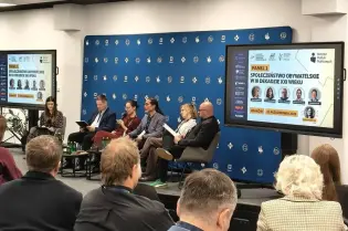 Zdjęcie przedstawia uczestników panelu dyskusyjnego siedzących na scenie przed niebieską ścianką. Na ekranach wyświetlono slajd z tytułem „Społeczeństwo obywatelskie w III dekadzie XXI wieku”. 