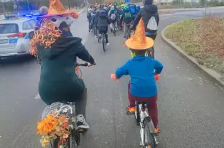 Grupa rowerzystów w kapeluszach, przejeżdżająca przez ulicę w jesiennej scenerii.