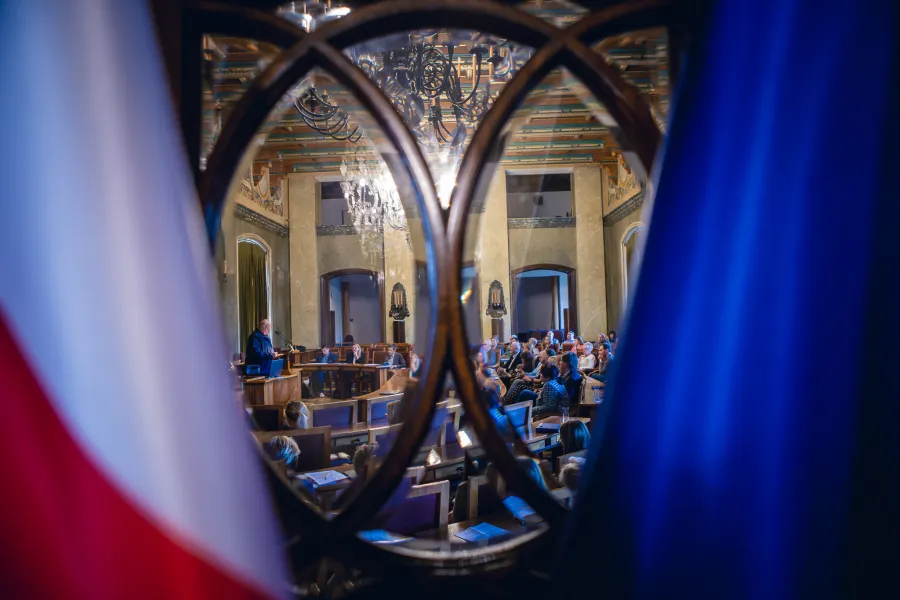 Widok na salę obrad z flagami w tle i mówcą na podium.