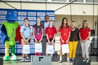 Uczestnicy zawodów sportowych z medalami i nagrodami, w tle maskotka i organizatorzy.