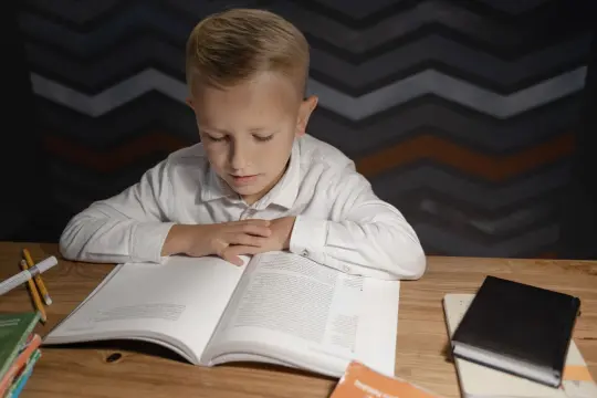 Chłopiec czytający książkę przy biurku z zeszytami i przyborami szkolnymi.