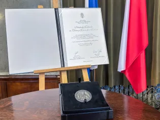 Dyplom z medalem na stole, flagi Polski i Unii Europejskiej w tle.