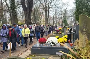 Grupa ludzi spacerujących po cmentarzu, otoczona nagrobkami i kwiatami.