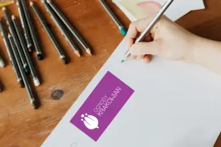 Osoba pisząca na białej kartce z logo organizacji w tle, otoczona przyborami do rysowania.