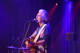 Mężczyzna grający na gitarze i śpiewający na scenie, oświetlony kolorowymi światłami.