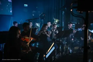 Muzycy grający na instrumentach w oświetlonym pomieszczeniu, z instrumentami dętymi i smyczkowymi.