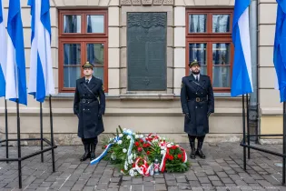 Dwaj żołnierze w mundurach stoją obok wieńca z kwiatów w barwach narodowych.